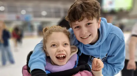 Dave Clarke Addy (left) sits in a chair and smiles, as he brother wraps his arm around her shoulders. 