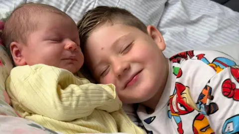 Ami Boyd Older brother Henry is smiling and lying next to Flossie, who is asleep on a bed wearing a yellow babygrow