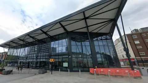 Google Sunderland train station. It is a tall glass building. There is a Tyne and Wear Metro sign in front of the entrance. Several people are walking past.