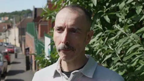 Matthew is standing on the pavement with a bush on the right-hand side of the picture and a street on the left. There are cars parked behind him. 