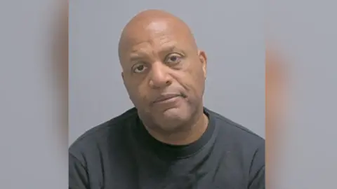 Norfolk Police Raymond Williams a man who is sitting down having his picture taken. He is bald and is wearing a black sweatshirt. He is looking directly at the camera.