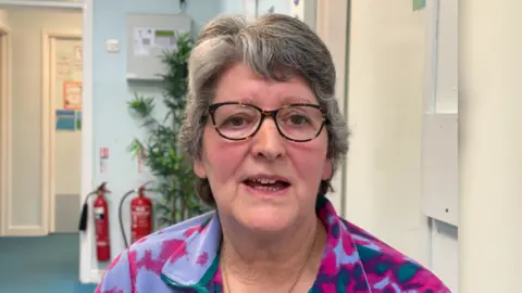 BBC A woman looks at the camera. She has short grey hair and glasses. She is wearing a top patterned with different shades of purple. A corridor with a potted plant is visible in the background. 