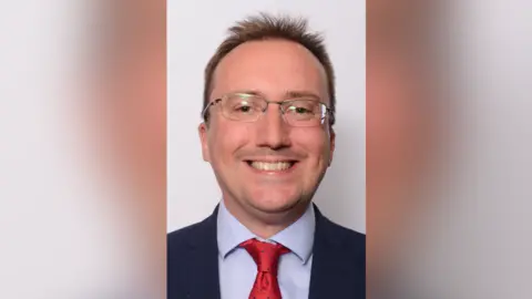 Keith Jones is pictured in a council portrait, wearing a red tie, blue shirt, black suit and glasses. He is in front of white backdrop.