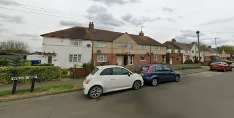 Google A row of blocks of two-storey houses. The nearest block has a brick ground floor and light-brown painted upper floor. There is a white car and blue car parked on the road outside the houses. There is a green hedge to the left of the picture. A white roadsign indicates WILLIS WAY in black lettering.