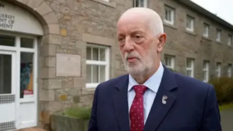 BBC Deputy Steve Luce is stood outside the environment department. He is wearing a navy blue blazer and red tie. 