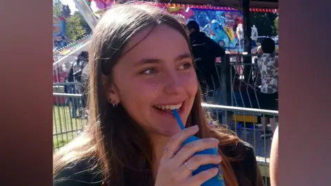 Chloe Longster stands next to a fairground ride sipping from a blue plastic cup through a straw. She is chuckling and looking to the right. She has long light brown straight hair and a small hoop earring.