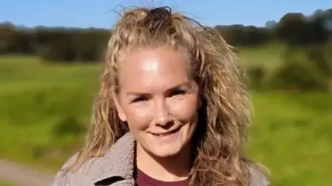 Family handout Carly smiles at the camera. She has thick curly blond hair and has a field and trees behind her