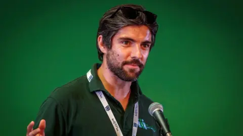 Born in Bradford A man with short brown hair speaks into a microphone he is wearing a green polo shirt and a white lanyard. He is standing against a green background