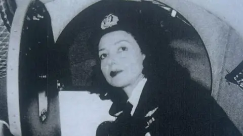 A black and white photo of Jackie Moggridge. She is wearing RAF uniform and is looking directly at the camera. She appears to be stood outside the cockpit of an aircraft. 