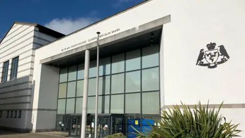 The outside of the courthouse, which is a large, white, glass-fronted building with the government crest on the right and Isle of Man Courts of Justice above the entrance.