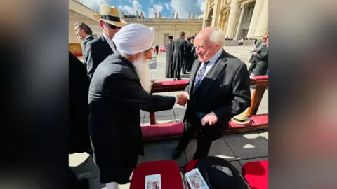 Bhai Sahib A bearded man, Professor Mohinder Singh, is shaking hands with a man, Irish President Michael D Higgin, who has white hair and is wearing a dark suit with a white shirt and a colourful tie.
