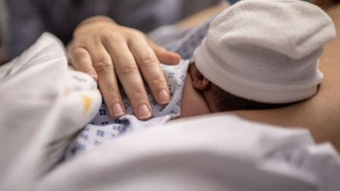A baby that's not long been born in hospital wearing a white hat and a woman's hand over its back