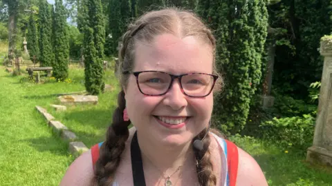 A woman is standing in an old graveyard. She had plaits in her hair and is wearing glasses. She is smiling at the camera. 