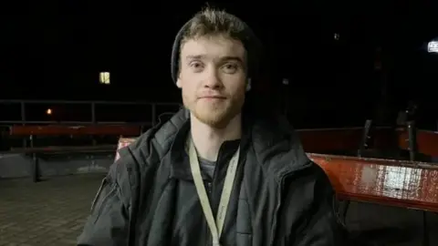 Chris Parry is sitting on a wooden bench in a pedestrian area in the dark. He has short blond hair and a goatee. He has a beanie hat pushed back on his head and a black anorak and has a lanyard around his neck.