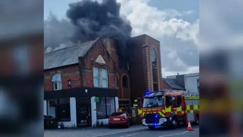 A brick building with large windows, with a thick cloud of smoke billowing out the top. A fire engine is stopped outside.