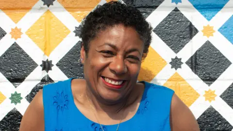Patrice Lawrence wearing a blue dress and smiling at the camera. She is sitting in front of a colourful wall.