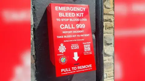 Avon and Somerset Police A bleed kit on a wall - it is red and has a lot of wording on it, including the sponsors and "emergency bleed kit to stop blood loss - call 999 - report incident - take bleed kit to patient