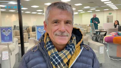 Spencer Stokes/BBC A man with a checked yellow and blue scarf, a blue puffer coat and grey hair. He is in a shopping centre smiling. 
