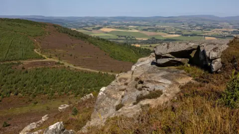 John Millard A view of the Rothbury Estate which has rocky outcrops overlooking a vast expanse of land. Below there is lots of green, brown and yellow fields and farmland with hills visible in the distance.