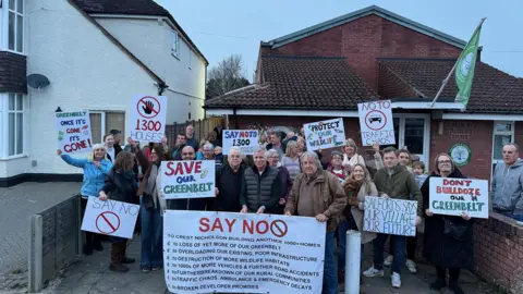 Handout Residents protest against the plans, standing outside houses with placards and banners. Slogans include "save our green belt" and "say no" and "don't bulldoze our green belt".