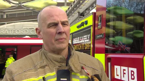 Graham Beers, a man wearing brown and yellow LFB uniform, looking at the reporter who is out of shot. A fire engine can be seen in the background