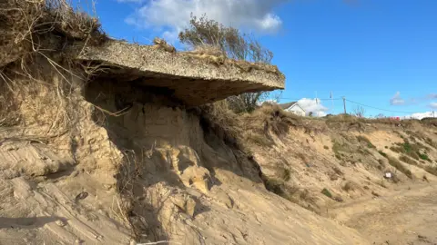 A view of a coastline that has been erroded away. A sand bank can be seen which some patches of grass and hedges. 