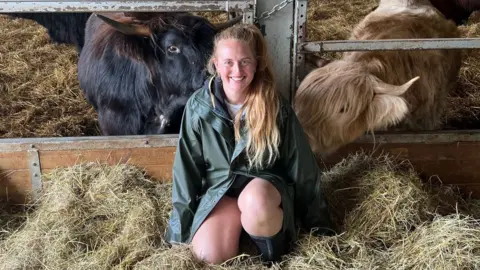 BBC Izzi Rainey - a blonde woman in her early 30s kneels in a barn in front of two of her cattle, one black and one blonde. Izzi s a while woman and her hair, tied in a pony tail, is over her shoulder reaching down to her middle. She wears a dark green rain mac and knee high boots and kneels amongst some hay - both cows are taking a keen interest in Izzi.