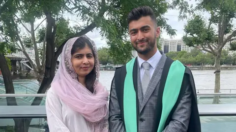 Khushal Yousafzai Malala, dressed in pink, with her brother Khushal Yousafzai as he graduates wearing black and light blue robes. They are standing on the London riverside, with green trees and river behind them.