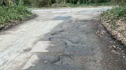 Contributed Ivy House Lane: a road edged by green verges. The left side of the road is light grey, the right side of the road is darker. The right side is pocked with holes and has been patched.
