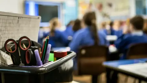 Pupils sit at desks in a classroom all facing forward and wearing a blue uniform.A plastic box with scissors, pend and pencils and a ruler are visible in the foreground wit the remainder of the picture blurred.