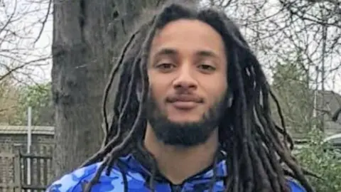 Sussex Police Nimroy Hendricks, who has a short beard and long dreadlocked hair and is wearing a tracksuit top with a blue camouflage design. He is stood in front of a tree, looking gently at the camera.