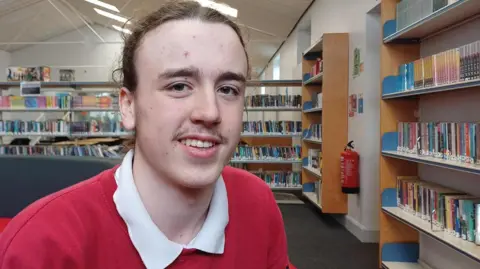 Debbie Tubby/BBC A teenage boy sitting in a library with shelves of books behind him and to the right. He has brown hair pulled back from his forehead, has a slight moustache and is smiling. He is wearing a red shirt with a red sweatshirt over it. 