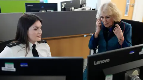 PA Queen Camilla sits next to a 999 call handler in the office of the Avon and Somerset Police Headquarters. She is sitting on an office chair and appears to be listening through a pair of headphones.