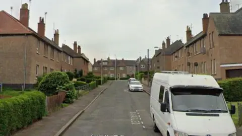 Google A white van is in the foreground. The street is lined with brown houses.