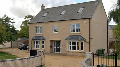 Front facing view of the house in Newtown Road, Ramsey. It has an off-road driveway with metal railings/gate and a brick wall at the entrance. There are three skylights in the roof, three windows on the middle floor of the house and two large bay windows on the ground floor. There is grass at either side of the house and trees. 