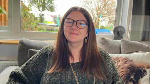 Amie Watson, sat on her sofa, looking into the camera. She is wearing an olive green cable knit jumper, and a long gold necklace. She has long brown hair tucked behind her ears, and a pair of tortoiseshell-framed glasses. She's sat on a grey sofa with a cushion with a picture of a Highland cow on it. Behind her is a large window, and you can see into the garden where there's a trampoline and a climbing frame. 