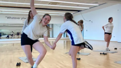 Two girls balance on one leg as they hold hands and smile in an exercise studio