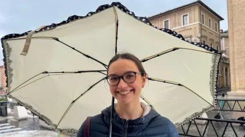 A young woman wearing glasses stands under an umbrella