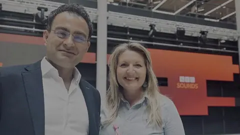 Surgeon Reza Arya stands next to Nicola Purdie in the BBC HQ in Cardiff. He has short, dark hair and wears glasses, a blue suit jacket and an open-necked white shirt. He is smiling. Nicola has long blonde hair. She is wearing a very light blue blouse. One large round drop earring can be seen in her left ear. They are in a studio with a red and orange panel behind them with the words "BBC Sounds" in BBC branding - three white squares containing the letters B,B,C in them in orange, and Sounds written underneath in light orange, all in capital letters.