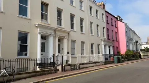 Row of three-storey Georgian type houses which are cream coloured with one pink house towards the back of the image. Part of a road and blue skies are also visible.