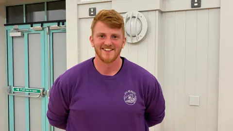 Topher has ginger hair and a ginger beard and wears a violent coloured top standing in the counting hall with a plain white wall behind him. 