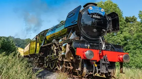 A dark coloured train with bright red bumpers in steam on a railway line