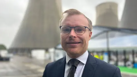 A ginger haired man wearing a suit and glasses stands in front of cooling towers.