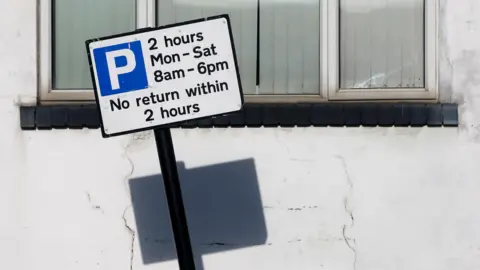 A white parking sign with a blue boxed in P, and a 2 hour limit. A white 