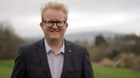 LDRS Independent Councillor for North Lisburn Gary Hynds smiling at the camera with a blurry landscape behind him. He has blonde hair, black framed glasses and is wearing an open neck shirt with a dark grey blazer.