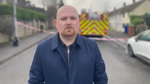 Alderman James Lawlor is looking into the camera. He's standing in the street. Emergency tape is up. A fire engine is parked. 