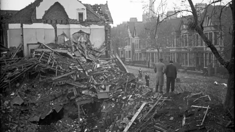 Mirrorpix via Getty Images Destroyed houses in Cardiff. Two men are standing on the rubble. The image is black and white