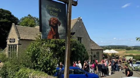 An historic pub under blue skies with rolling fields in the distance and a pub sign with a dog in the foreground. There is a queue of people outside.