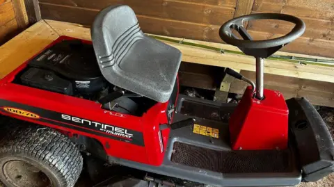 Fenland Orchards Project A red and grey ride-on mower parked in a wooden store. On the side are the words "Sentinel by Murray".