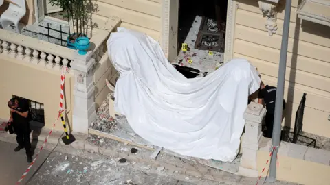 Frontage of a large cream coloured residential building in Monaco with damage to windows and a white sheet across the front steps and police standing near the front of the building. Photo: 30 June 2026